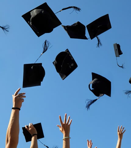 Graduation caps being tossed into the clear blue sky. Multiple hands are visible reaching upward, celebrating graduation.