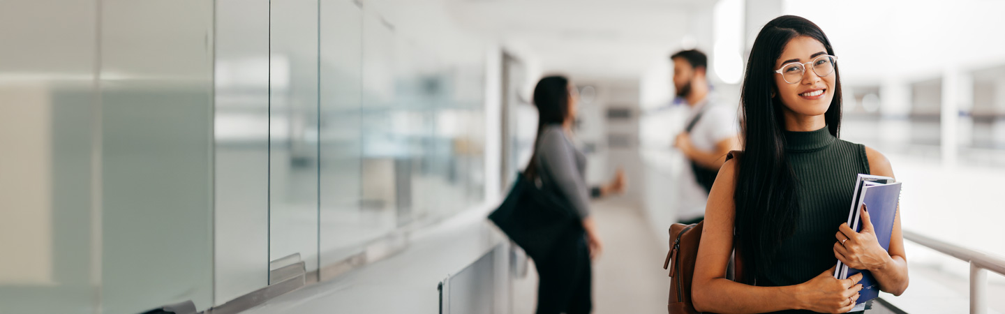A young woman with glasses, wearing a sleeveless top, holds books and a backpack in a bright hallway. She smiles, while two blurred people are in the background, one leaning against a glass wall.