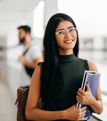 Smiling woman with long dark hair and glasses, wearing a sleeveless green top, holds notebooks and a shoulder bag. She stands in a bright, modern space, with a blurred figure in the background.