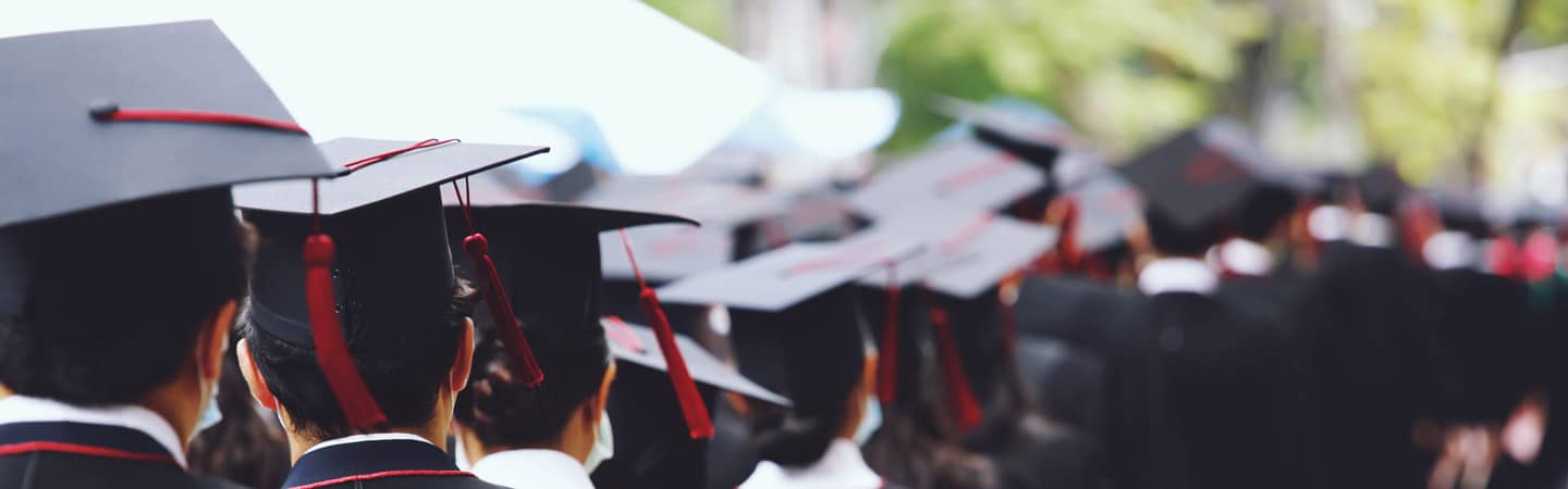 A group of students wearing black graduation caps with red tassels, walking in a line during a graduation ceremony. The background is blurred, showing trees and other graduates.