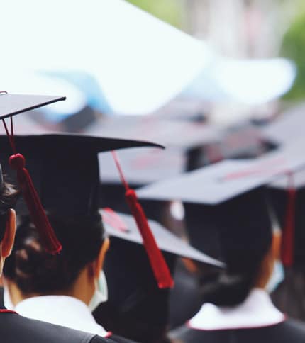 A group of graduates wearing black caps and gowns, with red tassels, stand in line facing forward. The background is blurred, suggesting an outdoor setting.
