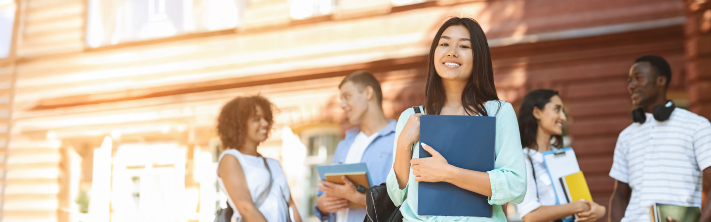 A group of students standing outdoors, smiling and holding books. One person is in the foreground, looking at the camera, while others chat in the background. The scene is bright and sunny, with a building visible behind them.