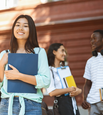 A young woman smiles and holds folders in front of a building. Two other people stand in the background, talking and holding notebooks. They all appear to be students.