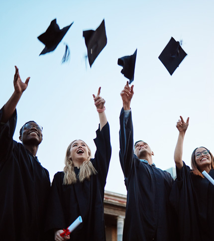 Four graduates in black gowns throw their caps into the air, celebrating against a clear sky. They hold diplomas and stand close together, smiling joyfully.