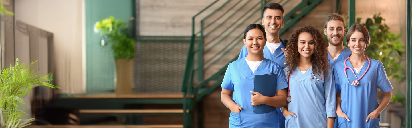 A diverse group of five healthcare professionals in blue scrubs stands together, smiling. One holds a folder, and another has a stethoscope. They are indoors, with plants and a staircase in the background.