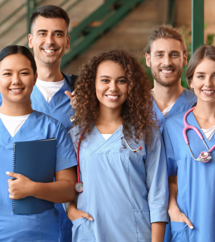 Five medical professionals in blue scrubs smile at the camera, some with stethoscopes and one holding a blue notebook. They stand indoors with green stairs and a plant in the background.