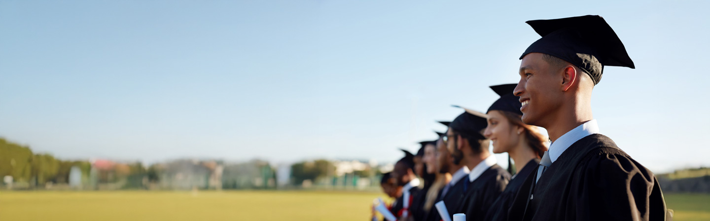 A line of diverse graduates wearing black caps and gowns stands in a grassy outdoor area on a sunny day. They are holding diplomas and looking forward with smiles, symbolizing achievement and new beginnings.