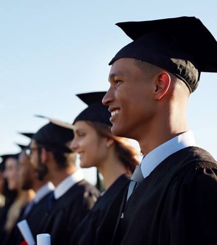 A group of graduates in black caps and gowns stand in a line outdoors. They are smiling and holding diplomas, with blue sky in the background. The focus is on a person in the foreground, showing a side profile.
