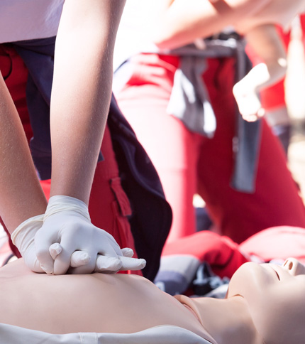 During a BLS Course on April 11, 2026, a person wearing gloves performs chest compressions on a CPR training mannequin, while others in red uniforms observe Basic Life Support techniques in the background.