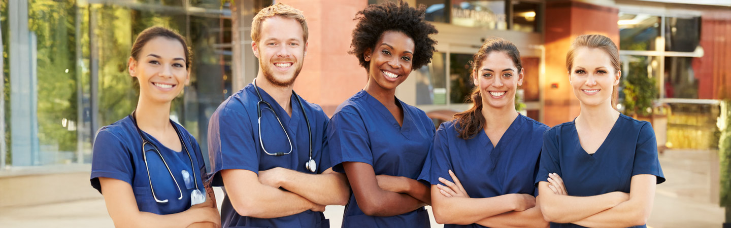 A group of five healthcare professionals in blue scrubs stands outdoors, smiling with arms crossed. There are two men and three women, showcasing diversity and teamwork. A medical building is visible in the background.