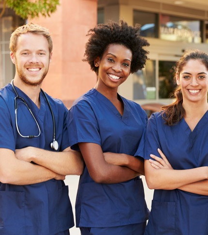 Three smiling healthcare professionals wearing blue scrubs stand with arms crossed outside a building. One man on the left has a stethoscope around his neck, and two women are next to him. They appear confident and approachable.