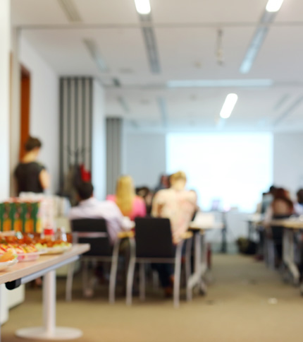 A blurred image of a conference room with people sitting on chairs facing a screen. A table with snacks or refreshments is visible in the foreground. The atmosphere appears to be professional and focused.