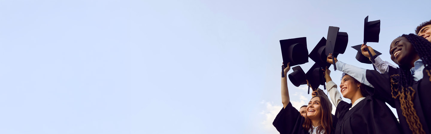 A group of graduates in black gowns cheerfully toss their mortarboards into the sky against a clear blue background.