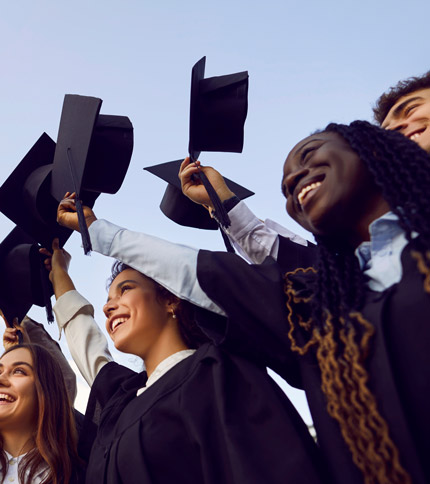 A group of diverse graduates in black gowns and caps celebrate together. They are outdoors, smiling, and holding their caps in the air against a clear blue sky.