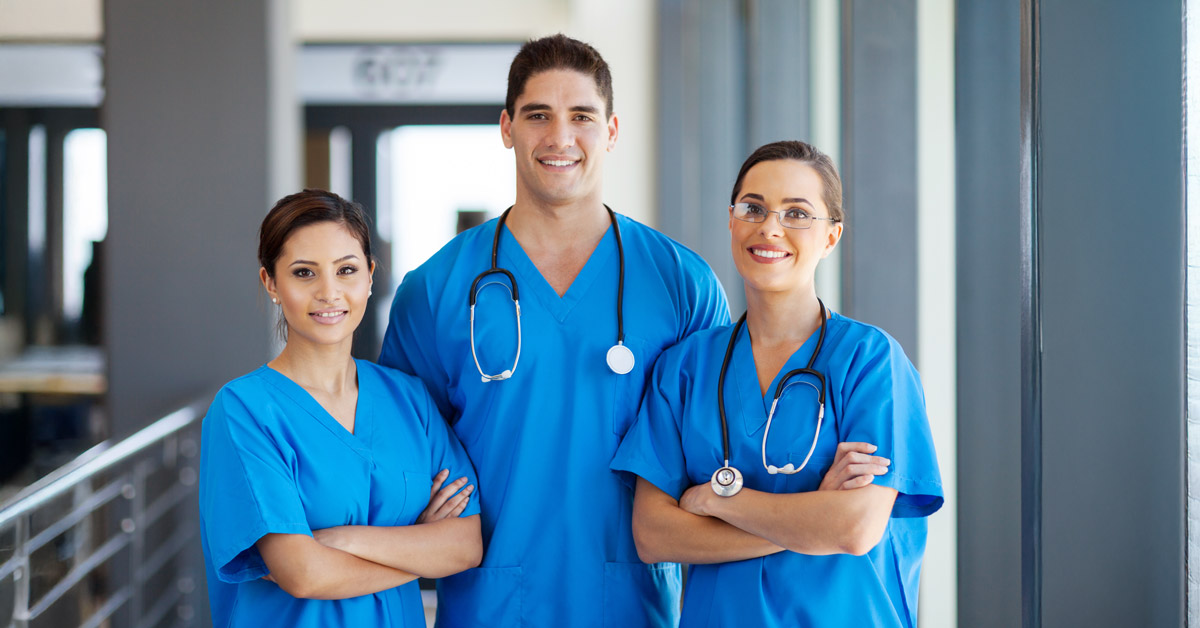 Three medical professionals in blue scrubs stand in a hallway, smiling at the camera. Two of them have stethoscopes around their necks. They stand in a well-lit environment, conveying a sense of confidence and professionalism.