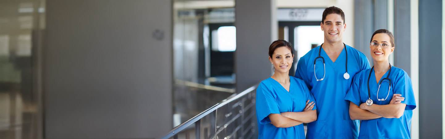 Three healthcare professionals in blue scrubs stand together, smiling and crossing their arms. They are in a modern, well-lit medical facility. Two have stethoscopes around their necks.