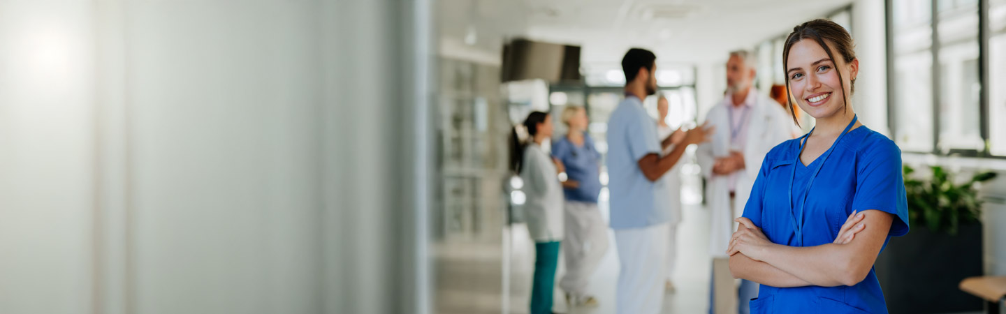 A smiling healthcare worker in blue scrubs stands with arms crossed in the foreground of a hospital corridor. In the background, several medical staff converse and review charts.