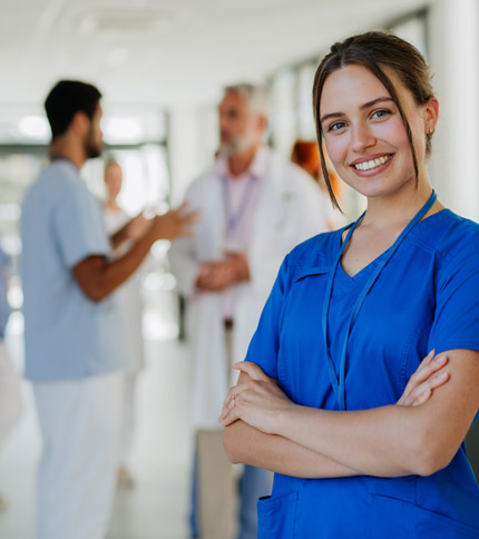 A smiling healthcare worker in blue scrubs stands with arms crossed in the foreground. In the background, medical professionals are engaged in conversation in a well-lit hallway.