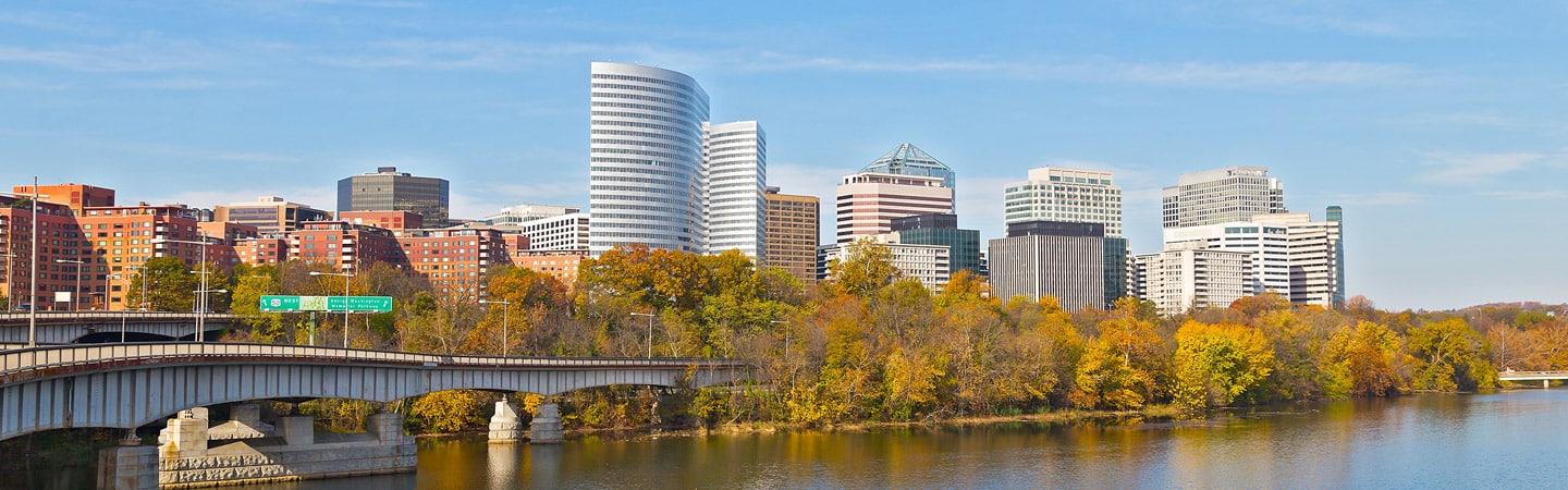 A cityscape with modern skyscrapers and a bridge over a river in the foreground. The trees along the riverbank display vibrant autumn colors under a clear blue sky.