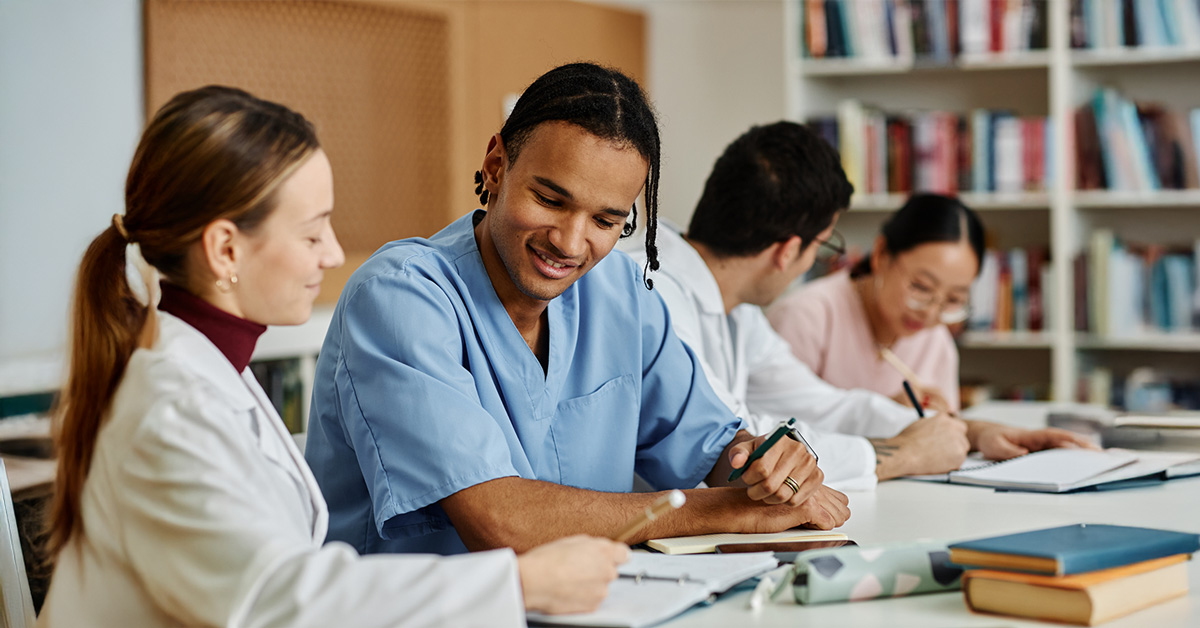 Four medical students study together at a table in a library. One wears blue scrubs, and the others wear white coats. Books and notebooks are open in front of them, with shelves of books in the background. They're engaged in discussion.