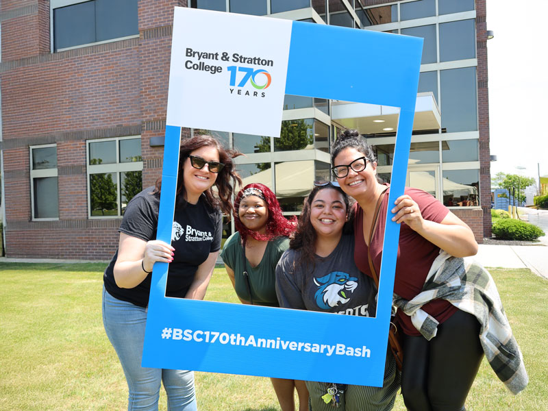 Four people smile while posing with a large blue frame outdoors. The frame reads "Bryant & Stratton College 170 Years" and "#BSC170thAnniversaryBash" at the bottom. A brick building with large windows is in the background.