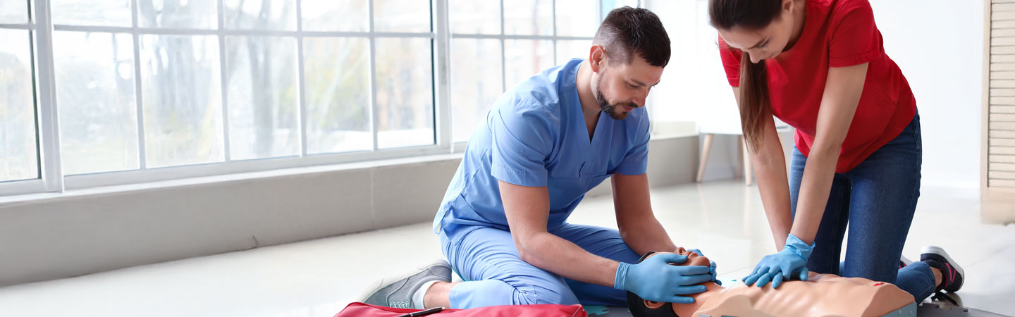 Two people in a medical training setting perform CPR on a dummy. One person in blue compresses the chest, while the other in red assists. They are positioned on a light-colored floor next to large windows.