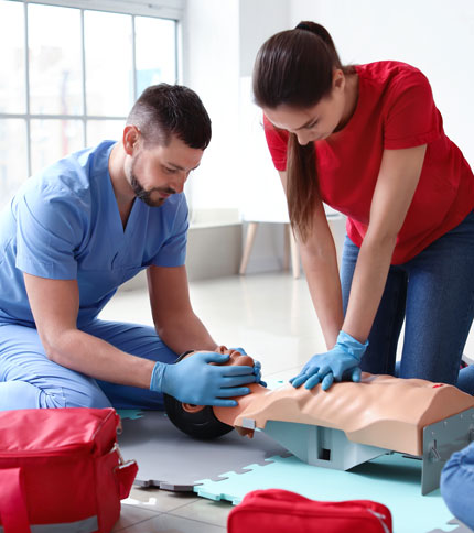 Two people are practicing CPR on a mannequin. One is wearing blue scrubs and gloves, giving ventilations, while the other in a red shirt performs chest compressions. Medical bags are on the floor nearby in a brightly lit room.