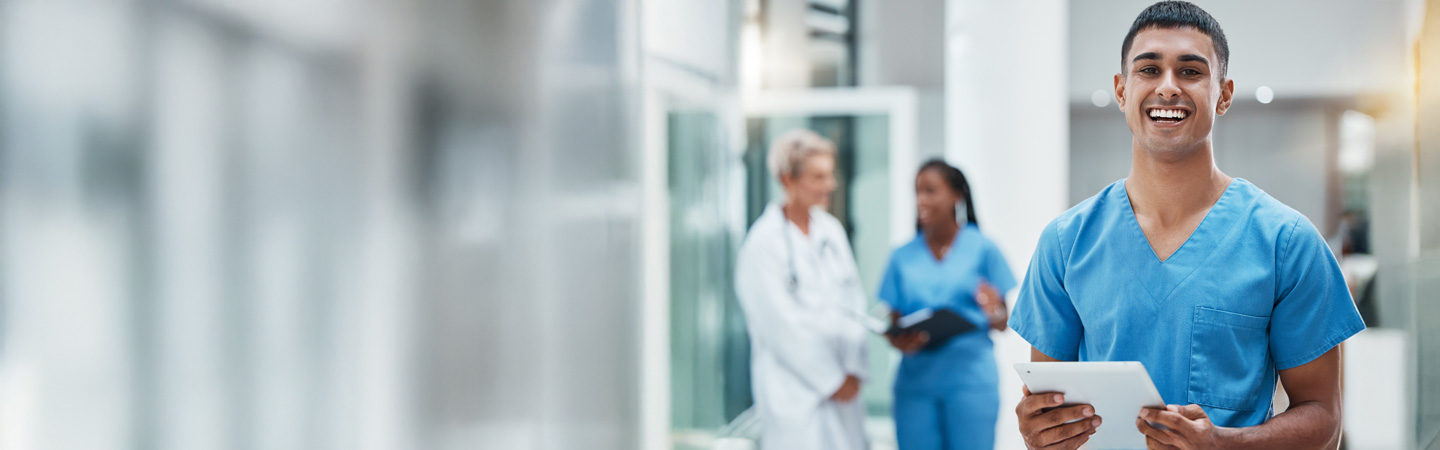 A smiling healthcare professional in blue scrubs holds a tablet in a bright, modern medical facility. Behind, two blurred figures in white coats converse, one writing on a clipboard, creating a busy hospital atmosphere.