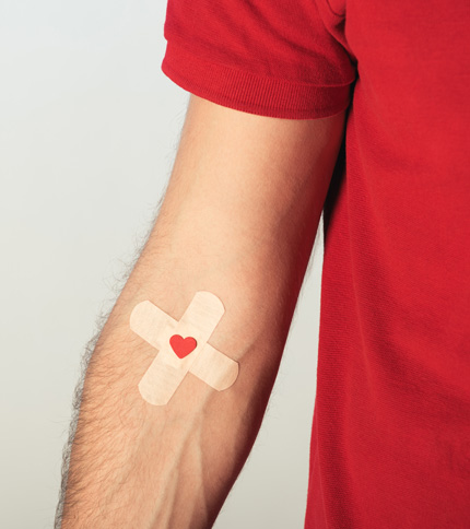 A person in a red shirt has two crossed adhesive bandages on their arm, forming an "X" shape. A small red heart is in the center of the bandages.