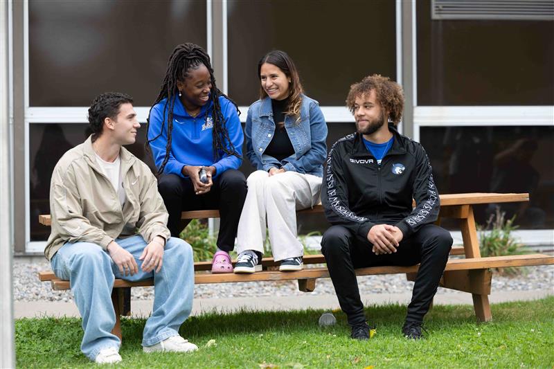 Four people sit on a picnic table and bench, smiling and talking. Two are seated on the table, and two on the bench. The background features windows and grass. They appear relaxed and engaged in conversation.