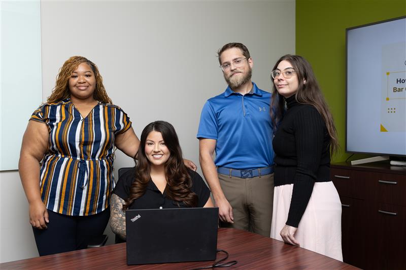 Four people are gathered around a table in a meeting room. One person is seated at a laptop while the others stand. There is a presentation screen in the background. They are all smiling and casually dressed.