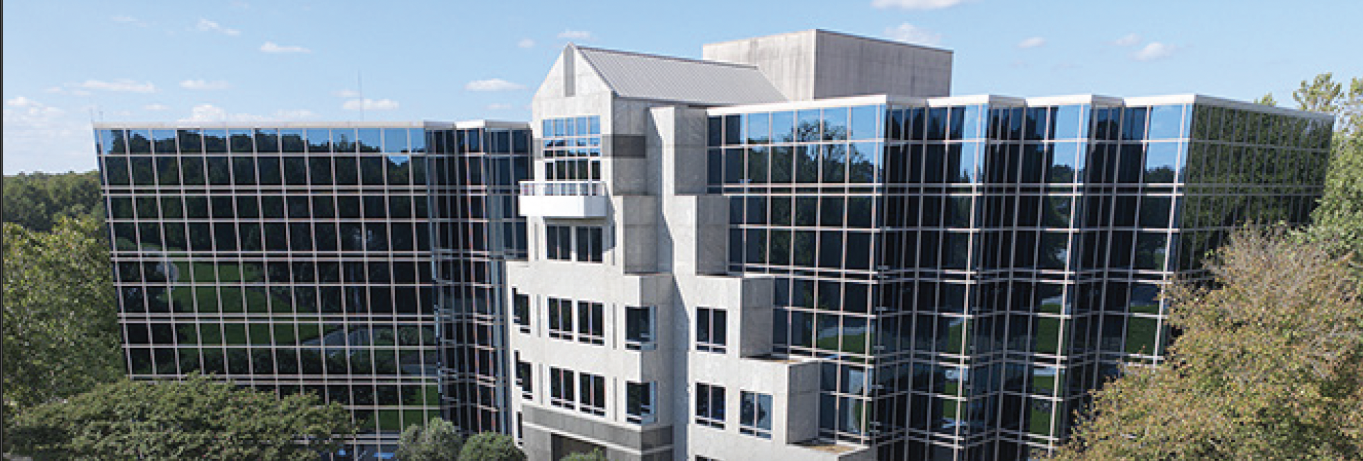 A modern office building with large, reflective glass windows and a central concrete structure, located in Upstate New York and surrounded by trees and greenery under a blue sky with scattered clouds.