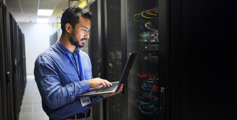 A person in a blue shirt stands in a server room, holding a laptop and looking at a server rack with cables and equipment. The surroundings are dimly lit with fluorescent lights above.