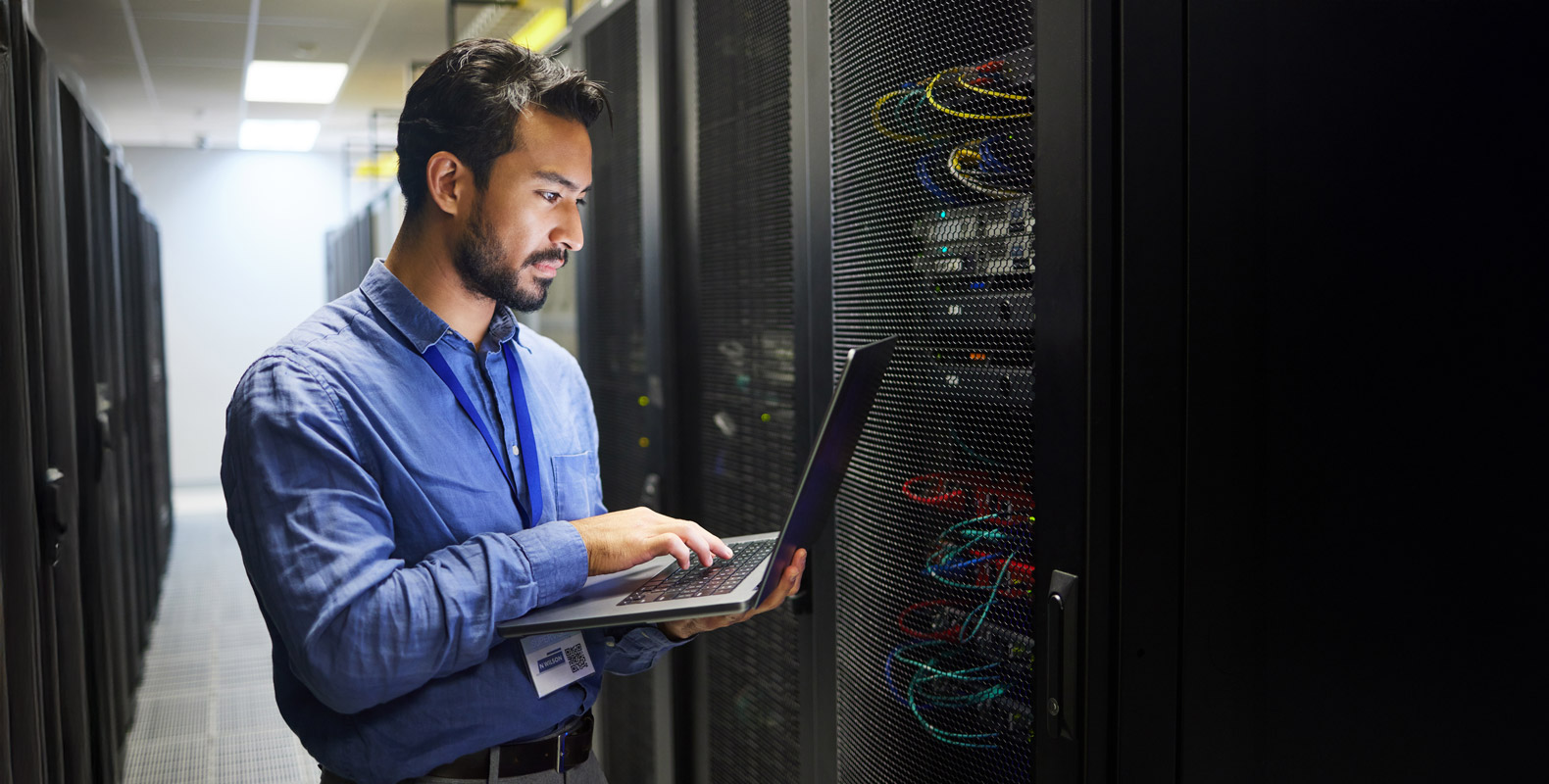 A person in a blue shirt stands in a server room, holding a laptop and looking at a server rack with cables and equipment. The surroundings are dimly lit with fluorescent lights above.