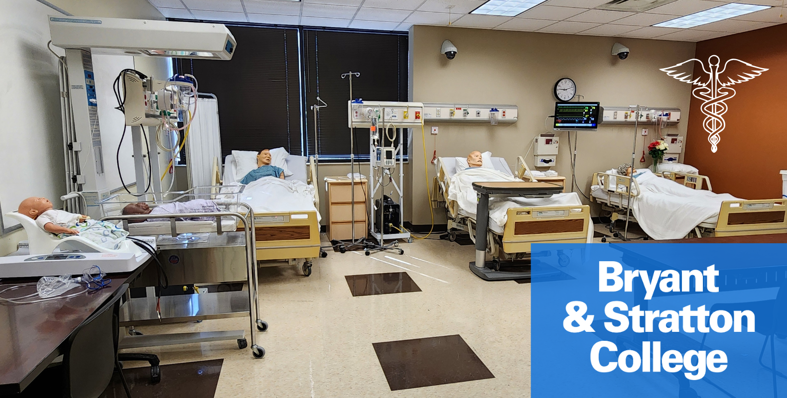 A simulated hospital room at Bryant & Stratton College features medical mannequins in hospital beds. Equipment like IV stands and monitors are present, with a caduceus symbol on the wall. The college's logo is visible in the corner.