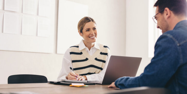 A woman is smiling and sitting at a table with a laptop and notebook, looking at a man seated across from her. They appear to be having a meeting in a bright room with white walls and paper documents on a board.