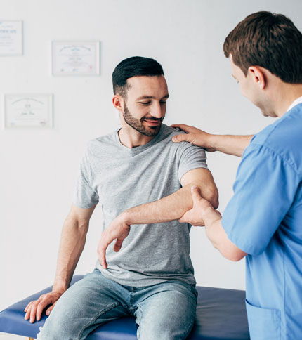 A man in a gray t-shirt is seated on a blue examination table, receiving a shoulder and arm check from a healthcare professional in blue scrubs, in a clinical setting with certificates on the wall.