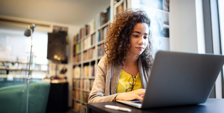 A person with curly hair in a yellow shirt and beige cardigan is sitting at a table using a laptop. They are focused on the screen. Bookshelves and a lamp are in the background, suggesting a library or study area.