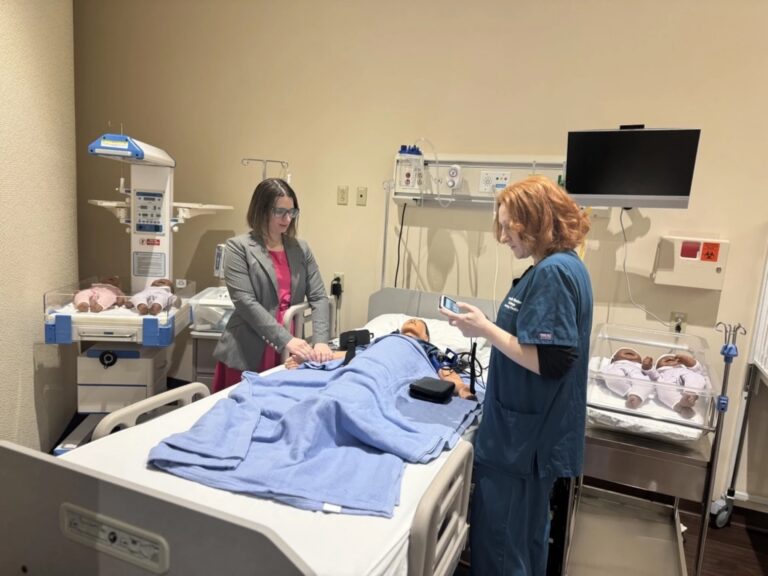In the Nursing Sim-Lab at Bryant & Stratton College in Greece, two women—one in scrubs and one in business attire—work with a patient simulator on a blue-covered bed, surrounded by medical equipment and baby mannequins.