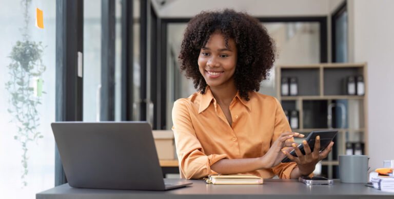 A woman sits at a desk with a laptop, smiling while holding a tablet. She is wearing an orange shirt and has curly hair. The workspace is modern, with shelves and large windows in the background.