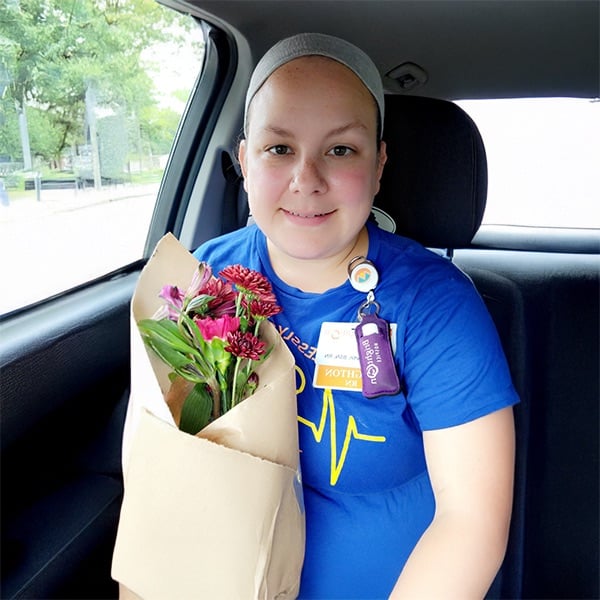 A smiling person wearing a blue shirt and a gray headband sits in a car holding a bouquet of flowers wrapped in brown paper. They have an ID badge and purple lanyard around their neck. Trees are visible through the car window.