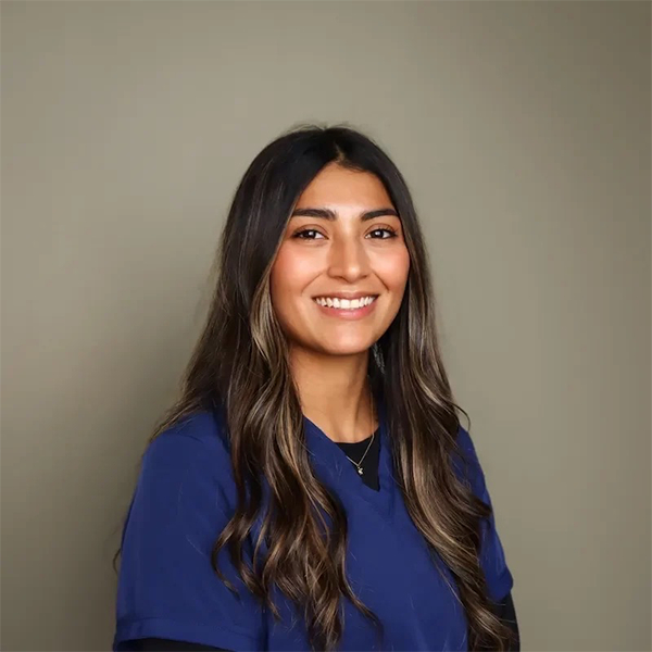 A woman with long, wavy brown hair smiles at the camera. She is wearing navy blue scrubs and standing in front of a plain, light olive background.