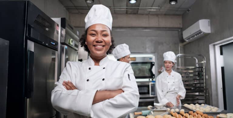 A smiling chef stands with arms crossed in a modern kitchen, wearing a white uniform and hat. Two other chefs work in the background, preparing food on a counter with pastries and baking trays.