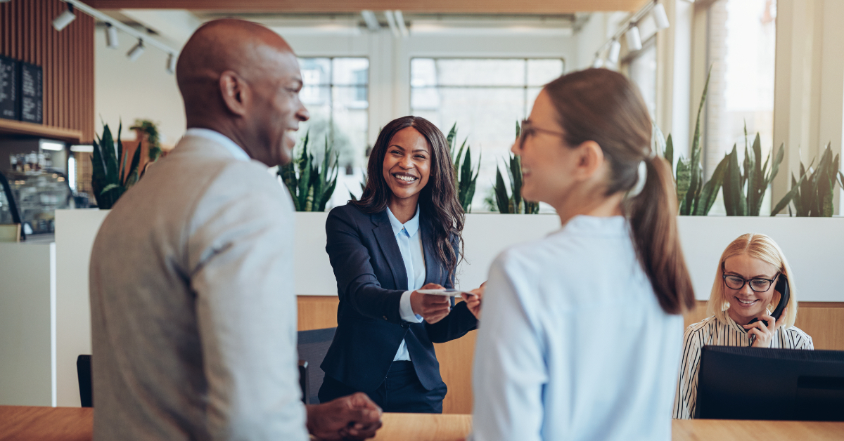 Three business professionals greet each other with smiles in a modern office; one woman hands a business card to a man while another woman works on a computer in the background.