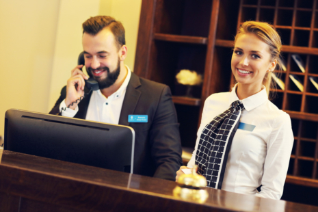 Two hotel receptionists stand behind a front desk; a man speaks on the phone and smiles at a computer, while a woman stands beside him, smiling at the camera. A bell and storage cubbies are visible.