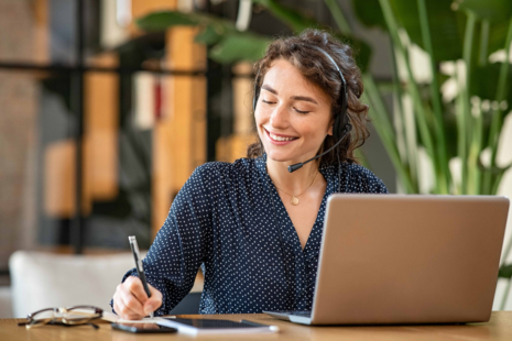 A smiling customer representative wearing a headset sits at a desk with a laptop, writing in a notebook. She appears to be working or in a video call, with glasses and a smartphone nearby, and large green plants in the background.