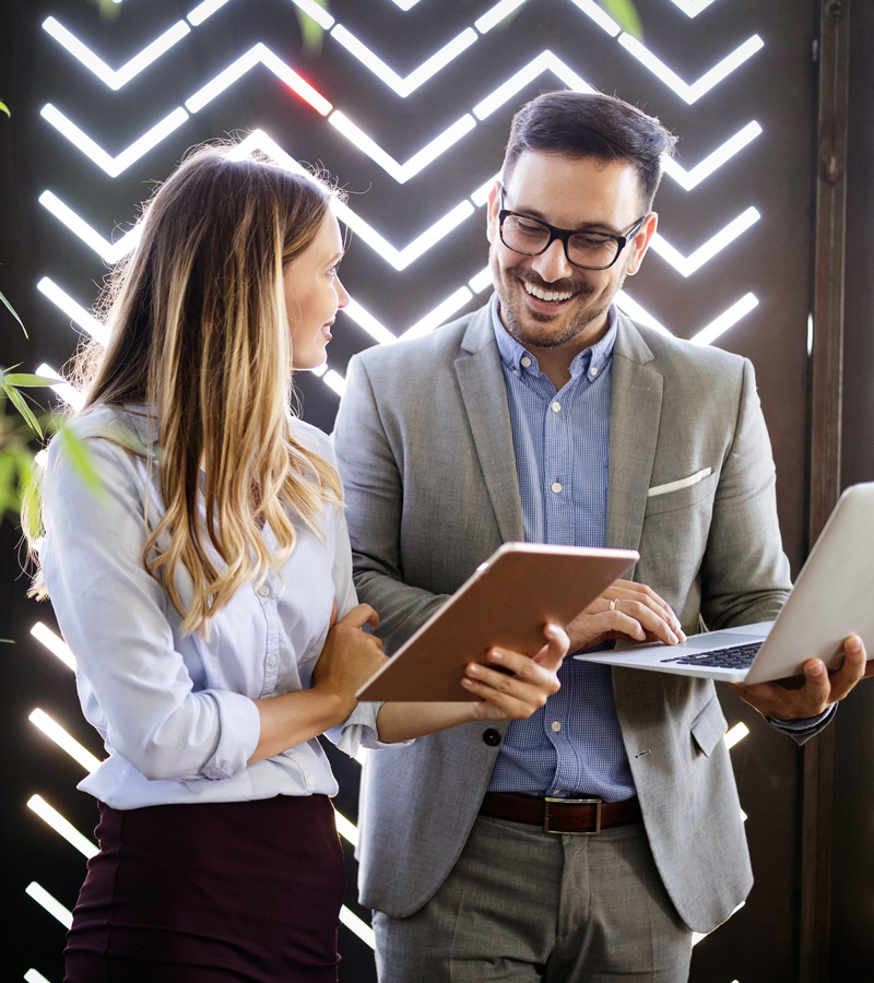 Two professionals, including a Customer Service Representative, stand and smile while discussing work—one holds a tablet, the other a laptop. They are in business attire with a patterned, light-filled wall in the background.