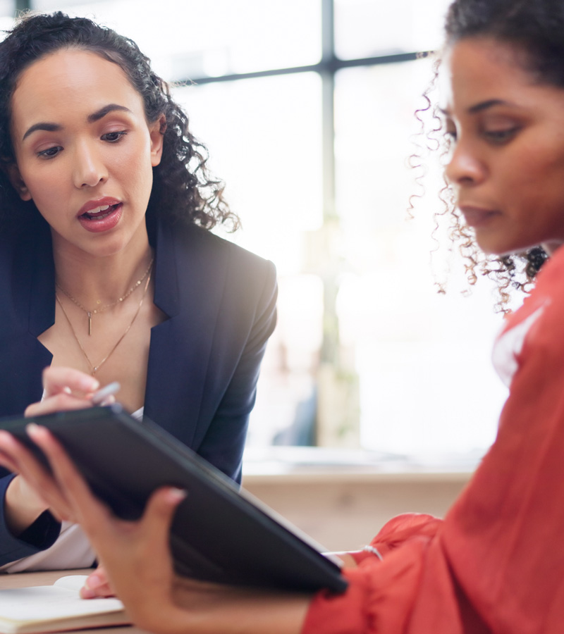 Two women sit together in a bright office—a Customer Service Representative in a blazer explains something while gesturing to a tablet held by the other woman, who is wearing a red top and listening attentively.