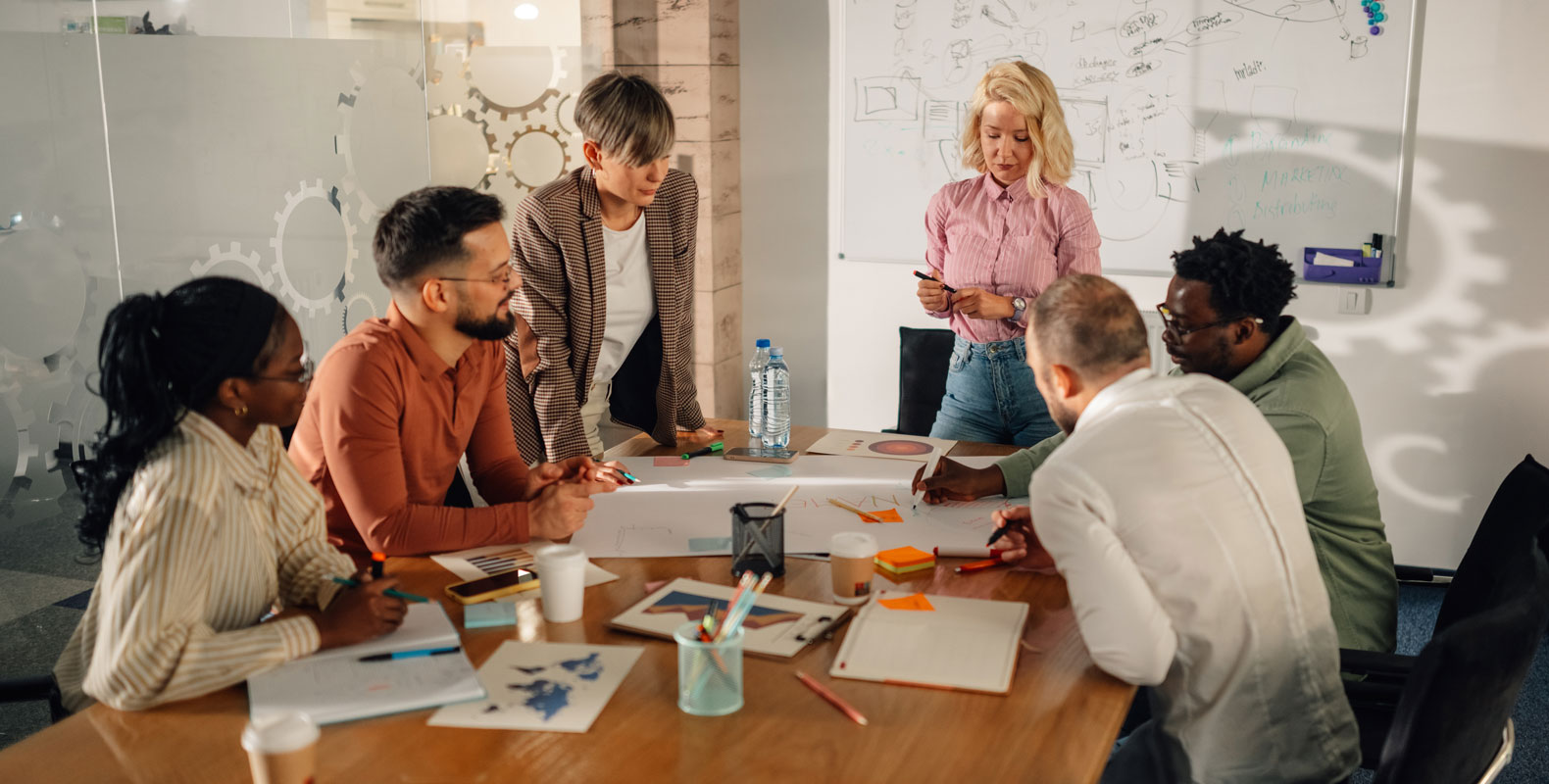 Six people are gathered around a conference table covered with papers and laptops, actively discussing a project. A woman stands and presents, while others listen and take notes in a modern office with whiteboards in the background.
