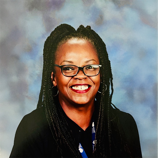 A woman with long braided hair, glasses, and hoop earrings is smiling widely. She is wearing a black top and a lanyard, with a soft blue and gray background behind her.